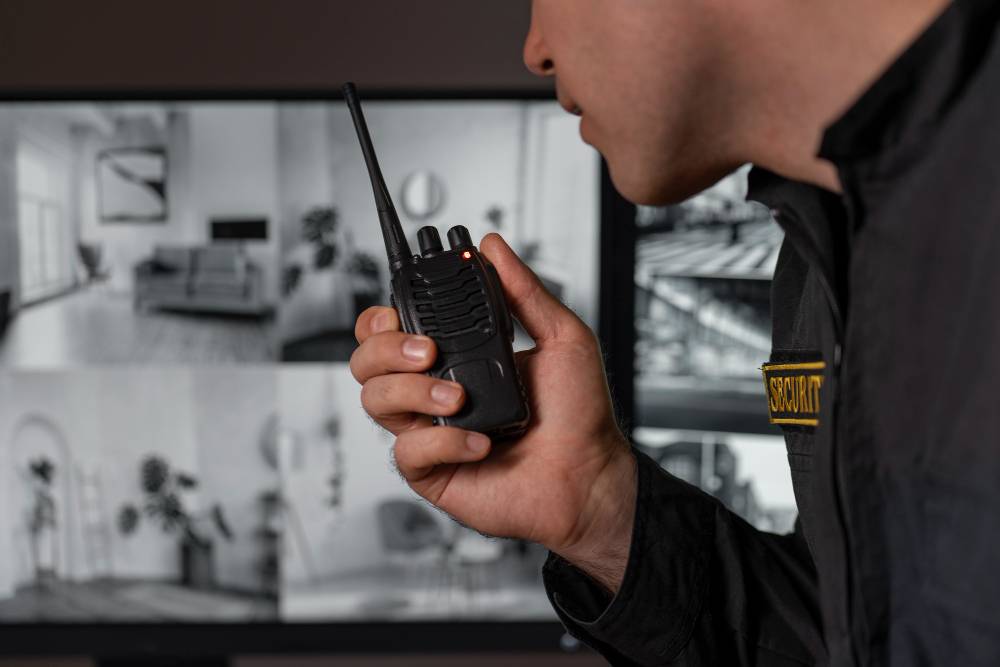Biztonsági őr rádión kommunikál a megfigyelő helyiségben, a háttérben kamerarendszer monitorai – élőerős őrzés és távfelügyelet.